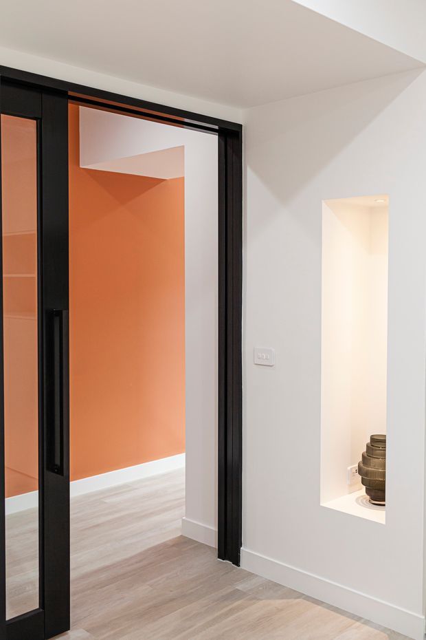 Interior view of a contemporary hallway or transitional circulation space, featuring a black-framed glazed sliding door opening to a room with a vivid terracotta-orange wall. The surrounding walls and ceiling are white, with pale timber-look flooring, a recessed illuminated display niche, and minimalist detailing.