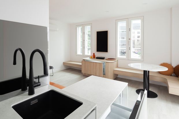 Bright open-plan contemporary kitchen and living area with a large pale island, black undermount sink and twin matte-black gooseneck tap fixtures in the foreground. Light timber built-in bench seating and cabinetry extend through the living zone, with a compact round dining table, wall-mounted television, and abundant white-framed windows creating an airy interior.