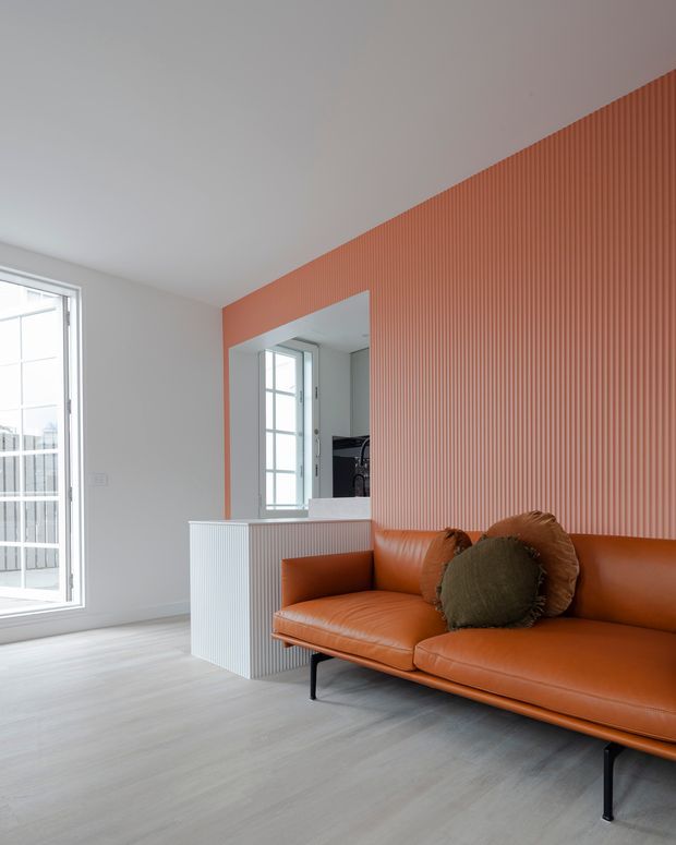 Bright contemporary living room with a long cognac leather sofa, black slim metal legs, and textured rust-orange vertical wall panelling. Light grey wood-look flooring, white walls and ceiling, and large multi-pane glazing create an airy, refined interior. A pass-through opening and ribbed built-in partition add architectural interest.