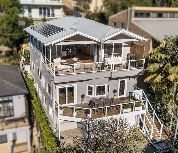 Elevated exterior view of a multi-storey coastal-style residence with a light grey weatherboard facade, white trim, multiple timber decks and balconies, broad roof overhangs, and a rooftop solar panel array. The home is surrounded by neighbouring houses and tropical landscaping, with outdoor seating visible on the upper terrace and stairs connecting the levels.