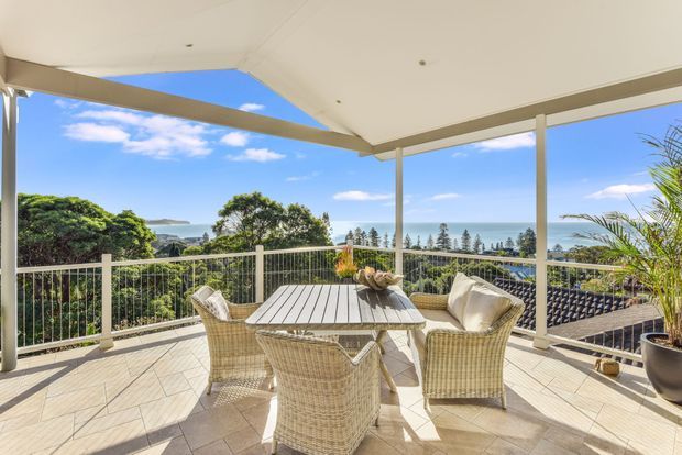 Bright covered outdoor terrace with a wicker dining table and chairs, overlooking dense greenery and a blue coastal or water vista. The space has a light tiled floor, slender pale structural posts, a flat covered roof, and open balustrade glazing, creating an airy elevated entertaining area.