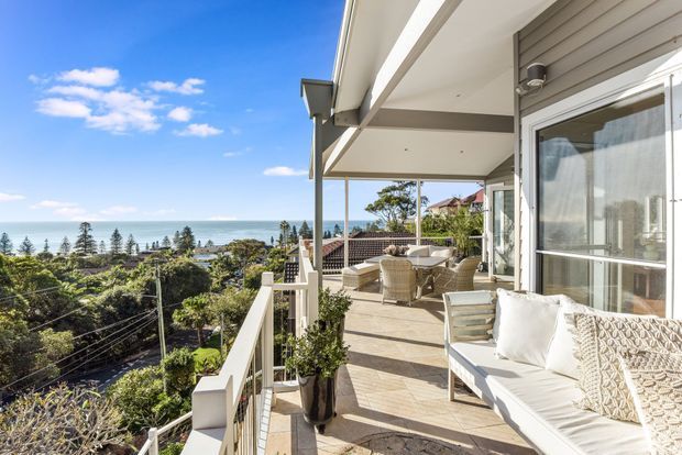 A bright, elevated outdoor terrace overlooking a coastal suburban landscape and the ocean. The balcony features a covered roof structure, white balustrades, pale tiled or stone-look flooring, a light outdoor sofa with textured cushions, wicker dining chairs, and a table. Large glazed sliding doors connect the terrace to the interior, while exposed beams and simple contemporary detailing define the architecture.