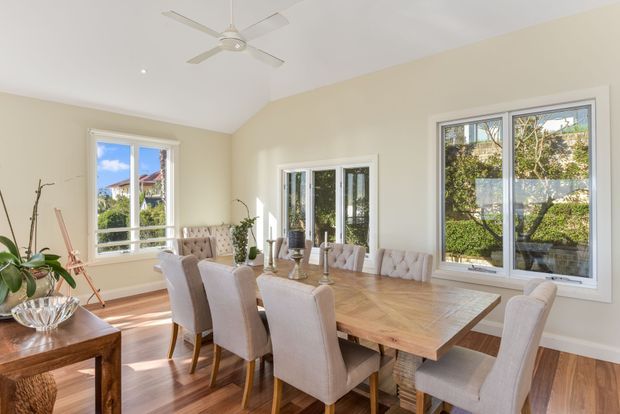 Bright, spacious dining room with a large rectangular wooden dining table surrounded by upholstered dining chairs. The room has cream walls, white-trimmed multi-pane windows and glazed double doors, polished warm timber flooring, potted plants, and a white ceiling fan. Natural light and garden views create an airy, inviting atmosphere.