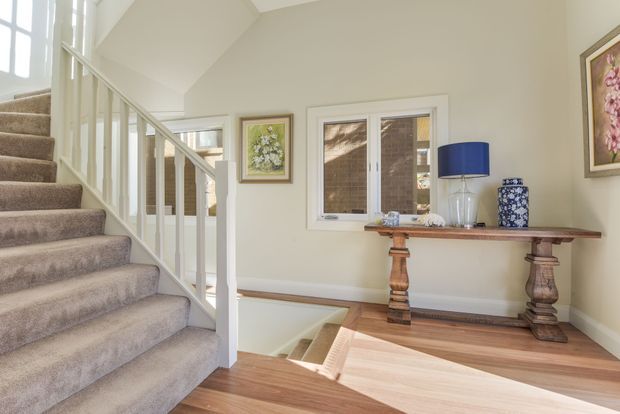 A bright, refined stairway and landing with a carpeted staircase, white-painted balustrade, pale neutral walls, and warm timber flooring. A traditional timber console table sits beneath a window, styled with a blue lamp, decorative vase, and framed botanical artwork. The stairwell has a high, angled ceiling and abundant natural light.