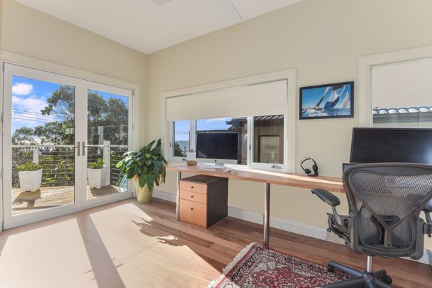 Bright home office with a built-in wall-to-wall desk beneath a wide interior window, an ergonomic office chair, framed artwork, indoor plant, and large glazed French doors opening to a timber deck. The room has warm beige walls, a white ceiling, timber flooring, and strong natural daylight.