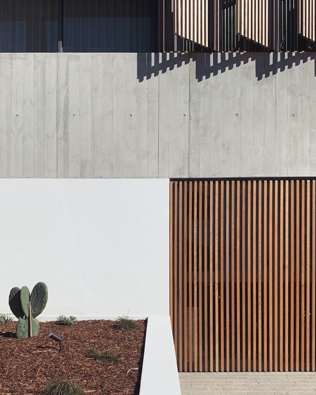 A close-up exterior view of a contemporary building facade featuring pale vertical board-formed concrete, smooth white rendered walls, dark glazing, and prominent vertical timber slatted screens. The slats cast strong geometric shadows across the concrete. A timber-slatted garage or entry door occupies the lower right, opening onto light grey rectangular paving, while a small landscaped garden bed with a cactus is visible at lower left.