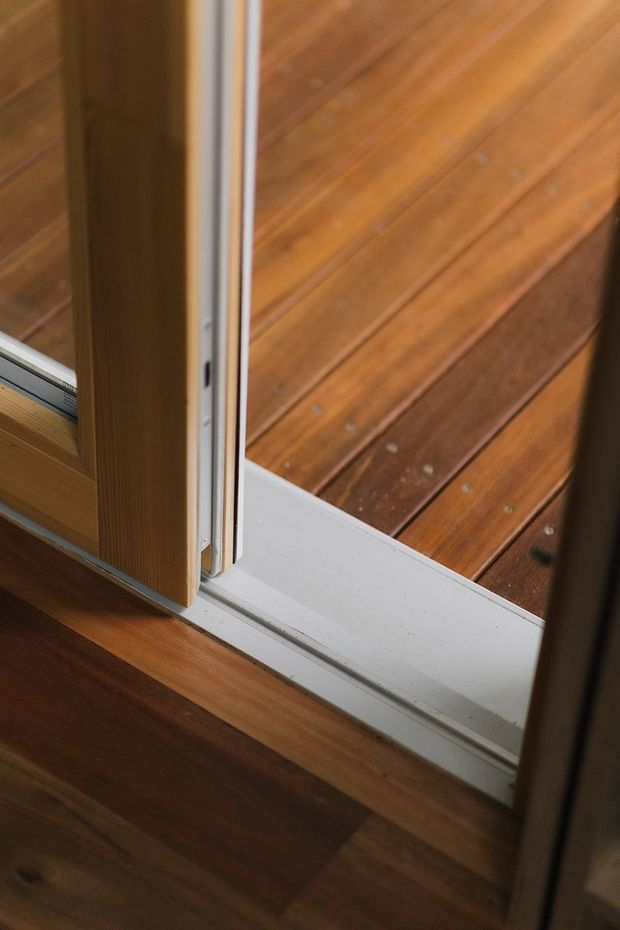 Close-up detail of a timber-framed glazed door or window opening onto a timber deck. The image focuses on the white sill and track, warm brown wood flooring inside, and horizontal outdoor decking beyond.