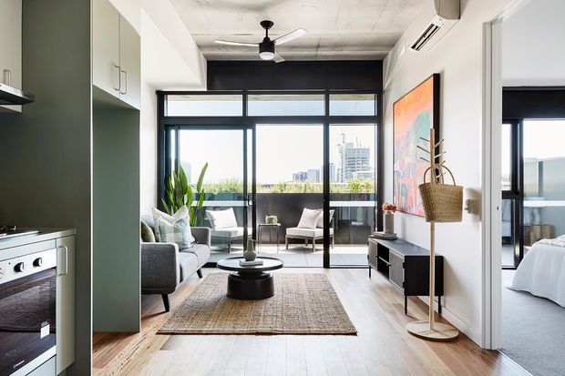 A contemporary open-plan apartment interior viewed from the kitchen toward a living area and balcony. The space features white cabinetry, pale timber flooring, a grey sofa, black-framed floor-to-ceiling glazing, a black accent wall, a compact round coffee table, and colourful artwork. A bedroom is visible through the doorway on the right. The ceiling has exposed concrete texture and a ceiling fan, with urban buildings visible beyond the balcony.