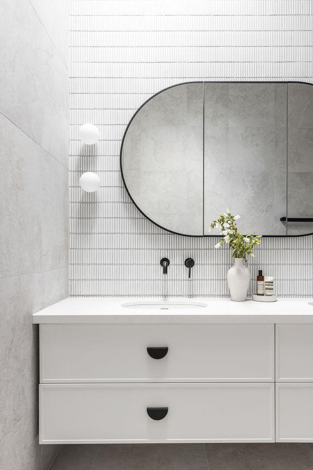 Contemporary bathroom vanity with a long floating light-grey cabinet, white integrated basin, pale stone-look benchtop, dark hardware, and an oval black-framed mirrored cabinet. The focal wall is clad in narrow vertically ribbed white tiles, with large-format grey wall and floor tiles creating a restrained monochromatic scheme. Spherical wall lights and a vase provide subtle decorative detail.