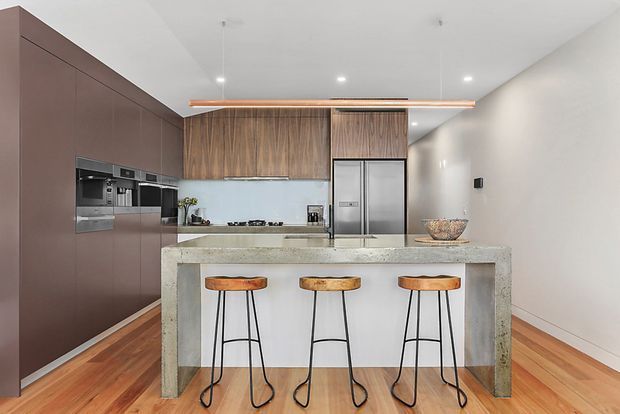 Contemporary open kitchen with a large concrete island and three slim bar stools. Full-height dark taupe cabinetry houses integrated appliances on the left, while warm timber-grain cabinetry lines the rear wall. Stainless-steel appliances, a pale benchtop, timber flooring, white walls and recessed ceiling lights create a clean, warm, modern interior.