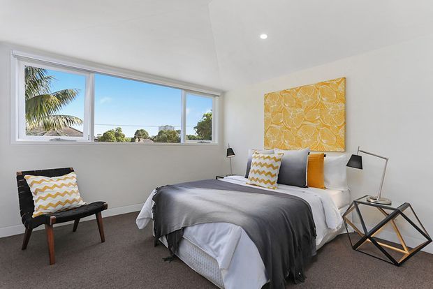 Bright contemporary bedroom with a large horizontal window overlooking trees and a palm, a neatly made bed layered in white, grey, and mustard textiles, a black accent chair, bedside lamps, and a geometric side table. White walls and ceiling create an airy setting, while the mustard patterned artwork and cushions add warmth and contrast.