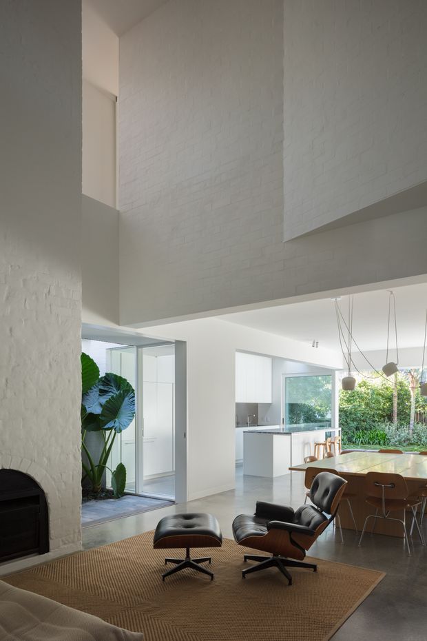 A bright, open-plan contemporary interior with a double-height white-painted brick wall and mezzanine-like upper volume. The space combines a living area with a fireplace, woven rug, leather lounge chair and ottoman, a dining area with pendant lights, and a minimalist white kitchen opening toward a leafy courtyard through large glazed doors.
