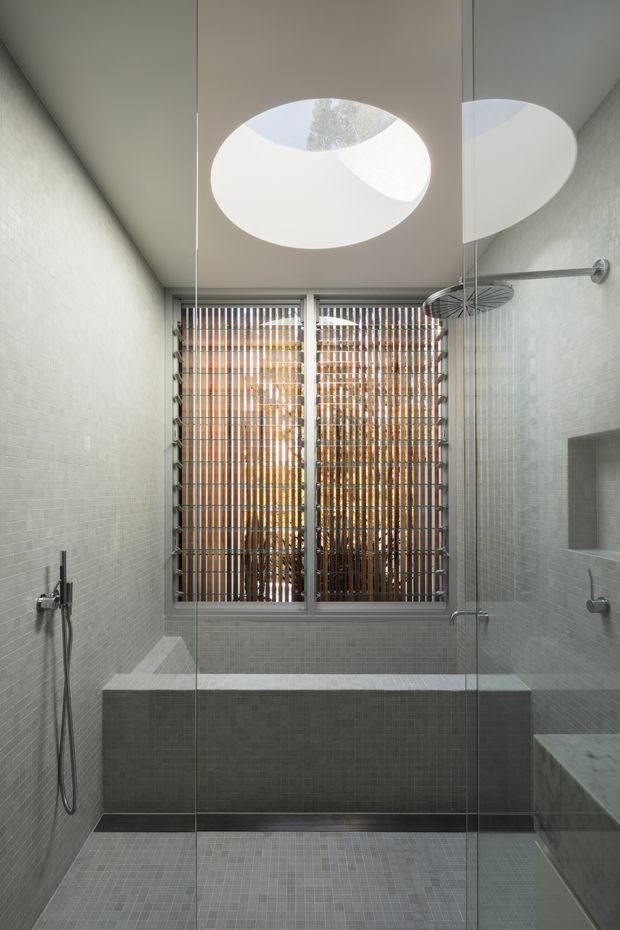 Contemporary bathroom with a fully tiled walk-in shower and bathtub zone enclosed by clear glass panels. Small pale grey mosaic tiles cover the walls, floor, and built-in tub surround. A large circular skylight brings in natural light, while a high window features timber vertical screening and louvres. Brushed metal shower fittings and a marble-clad vanity edge are visible.