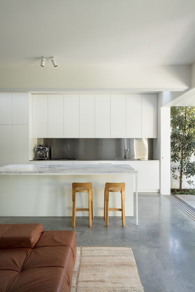 A bright, minimalist open-plan kitchen with full-height white cabinetry, a stainless-steel splashback, pale stone island benchtop, and two timber stools. The kitchen opens directly to an outdoor area through wide glazing or folding doors. A polished grey concrete floor, brown leather sofa, and neutral rug are visible in the foreground.
