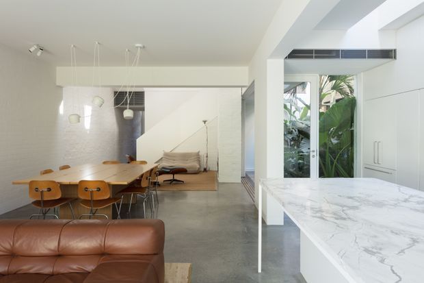 Bright open-plan contemporary interior combining a kitchen, dining area and living space. A large white marble island dominates the foreground, while a timber dining table with brown leather chairs and a brown leather sofa occupy the adjoining areas. White walls and built-in cabinetry, polished concrete flooring, pendant lighting, and full-height glazing to lush greenery create a clean, airy space.