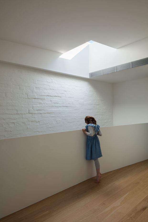 A minimalist interior corridor or landing with a white-painted exposed brick wall, pale timber flooring, and a half-height partition. A large angular skylight brings natural light into the space, while a linear ventilation grille runs along the upper wall. A child stands at the partition, looking into an unseen area.