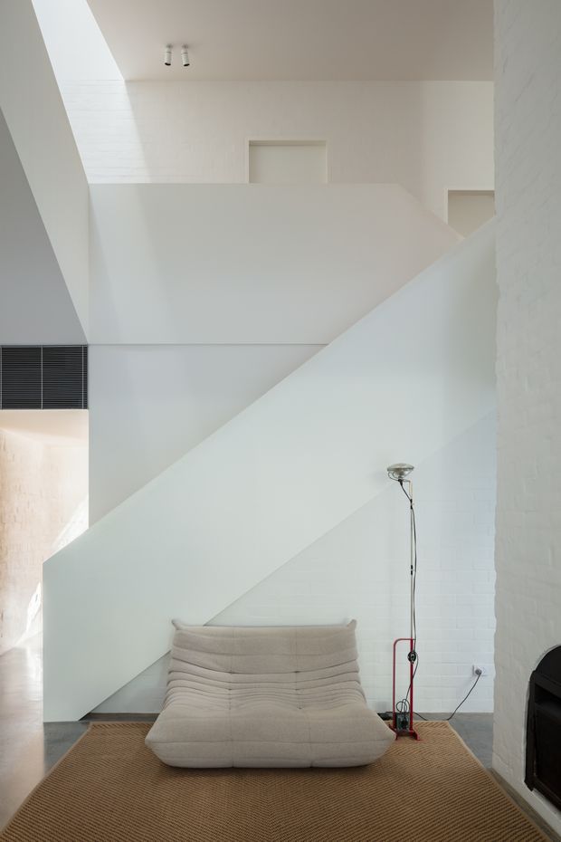 A minimalist white interior centered on a sculptural staircase with solid geometric balustrades and exposed white-painted brick walls. A low cream lounge chair sits on a large natural brown woven rug, beside a slender floor lamp. A dark fireplace is partially visible at the right edge, and a high-level vent and recessed openings emphasize the tall, airy volume.