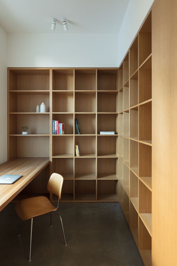 Compact home office lined with floor-to-ceiling built-in timber shelving and open cubby storage on three sides. A timber desk extends from the shelving, paired with a simple wooden chair. The room has white walls and ceiling, dark polished-looking flooring, minimal ceiling spotlights, and sparse books and decor.