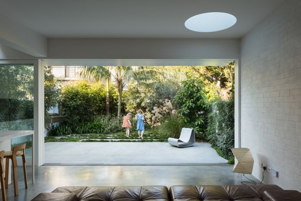 A bright contemporary living room opens directly onto a lush private garden through expansive full-width glazing and sliding doors. The room features a polished concrete floor, white brick wall, broad white ceiling, circular skylight, low dark leather seating, and a sculptural white lounge chair. A dining area is partially visible at left, while two people stand in the garden beyond.