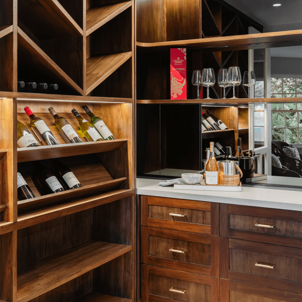A richly detailed built-in home bar and wine-storage area with dark timber cabinetry, angled wine racks, open shelving, glassware, wine bottles, and a pale stone benchtop. Warm integrated lighting highlights the joinery, while a window and mirror-like reflective surfaces add depth.