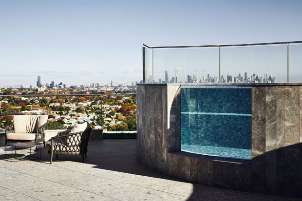A rooftop terrace with a compact elevated swimming pool or spa clad in dark stone, featuring a vivid blue tiled interior and clear glass balustrades. Outdoor lounge chairs and a small table occupy the left side, while a dense suburban cityscape and high-rise skyline extend across the background. The scene is sunlit, contemporary, and panoramic.
