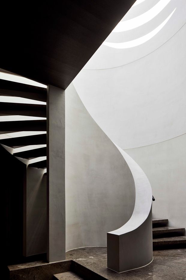 A dramatic minimalist interior focused on a sculptural curved staircase or ramp. Smooth pale plaster/concrete walls form a sweeping spiral enclosure, with narrow skylight openings casting bright bands of light from above. Dark stair treads and ceiling planes create strong geometric contrast against the light surfaces, while the rough dark floor reinforces the monolithic architectural character.