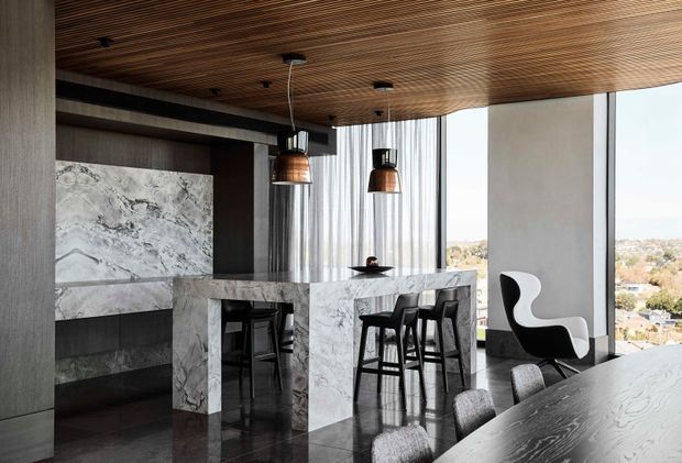 Sophisticated open-plan kitchen and dining area with dramatic white-veined marble island and fireplace surround, dark timber cabinetry and wall finishes, a richly slatted timber ceiling, pendant lighting, and floor-to-ceiling windows overlooking an urban landscape. Black bar stools and sculptural dining chairs create a refined, high-contrast interior.