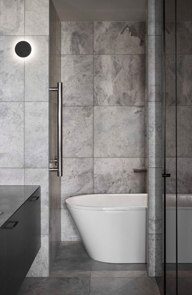 A sophisticated contemporary bathroom featuring large-format grey veined stone-look tiles across the walls and floor, a freestanding white bathtub, dark built-in vanity cabinetry, and a frameless glass shower enclosure. Minimal cylindrical wall lighting and brushed metal hardware add a refined, modern finish.