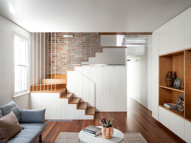 Bright contemporary living area with a grey sofa, round coffee table and built-in white cabinetry with warm timber display niches. A compact timber staircase with white vertical panelling rises beside an exposed brick wall, creating a strong architectural feature. Dark timber flooring, white walls and ceiling, and abundant natural light give the room a refined, warm feel.