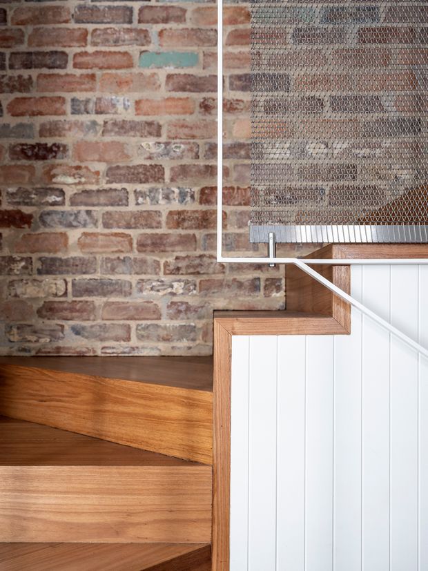 Close-up architectural detail of an interior staircase. Warm timber stair treads and risers meet a white vertically panelled stair balustrade, with a slim white metal handrail and wire-mesh guard above. An exposed, weathered red-brick wall forms the backdrop, creating a strong contrast between industrial masonry and refined timber joinery.