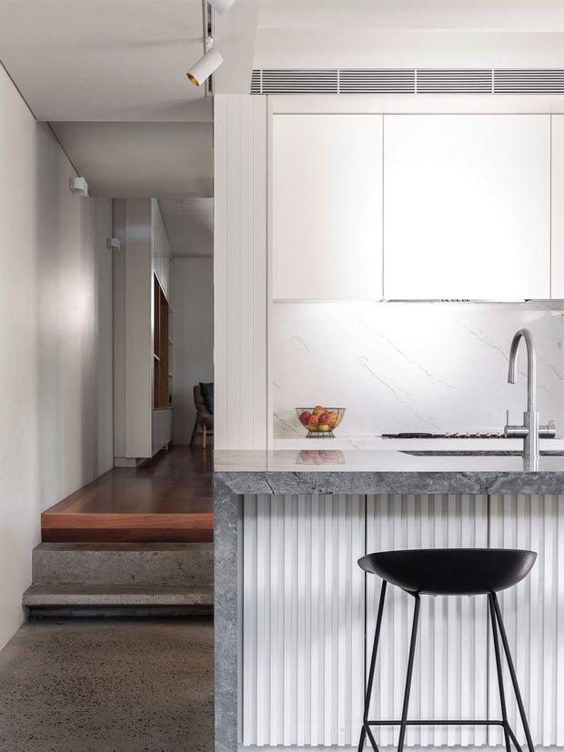 A bright, compact contemporary kitchen viewed beside a short set of concrete steps leading to a hallway. The kitchen features white flat-panel cabinetry, a white veined stone benchtop and splashback, a grey stone island edge, a vertically fluted white island front, stainless-steel tapware, and a black sculptural bar stool. Track lighting and a linear ceiling vent add crisp modern detailing.