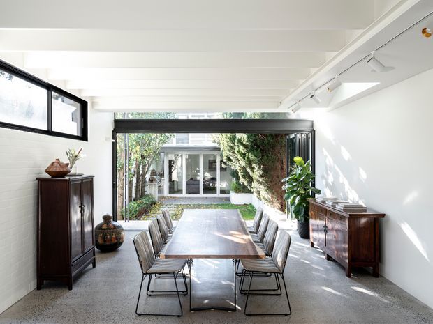 Bright, elongated dining room with a long rectangular table and matching upholstered dining chairs, opening through large glazed doors to a planted courtyard. The room has white painted brick walls, exposed white ceiling beams, polished concrete or terrazzo-like flooring, track lighting, dark timber storage cabinets, and abundant natural light. The overall feel is refined, airy, and contemporary with traditional furniture accents.