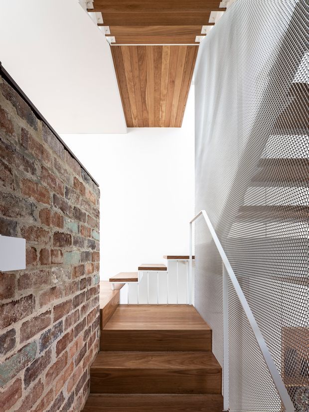 A bright, narrow interior stairway with rich timber stair treads and matching timber ceiling lining, bordered by an exposed reclaimed brick wall on one side and a perforated metal mesh balustrade on the other. White walls, cabinetry, and trim create a crisp contrast, while the stair rises toward a compact landing and upper-level circulation area.