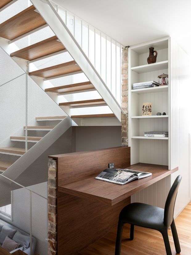 Bright contemporary stairway with open-riser timber stairs, white metal balustrades and perforated mesh screening. A built-in dark timber study desk with shelving occupies the space beside the stairs, with exposed brick detailing and pale vertical wall panelling. The composition highlights an efficient home office nook integrated into the stairwell.