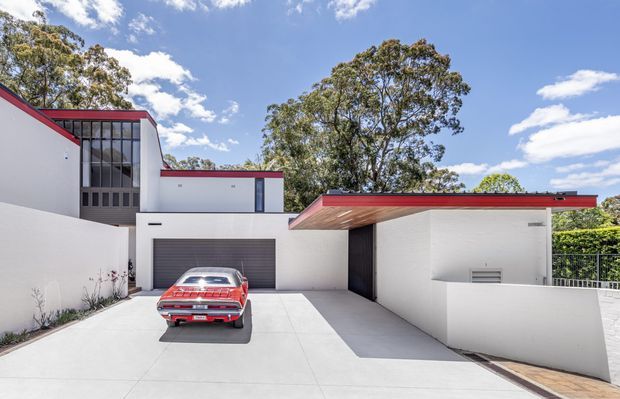 Exterior view of a contemporary white multi-level residence with a flat roof and bold red roof-edge trims. The image shows a broad concrete driveway, enclosed garage with a dark roller door, covered carport or entry canopy, tall black-framed glazing, and a small portion of a metal balcony railing. A vintage red car is parked in the driveway, with mature trees visible beyond the property.
