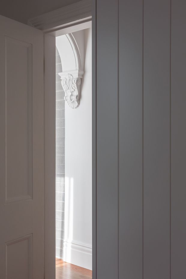 A softly lit interior view through a partially open paneled door into a refined hallway or adjoining room. The foreground features tall white vertical wall panelling or cabinetry, while the doorway reveals white decorative mouldings, an ornate carved corbel, a curved arch detail, white brickwork, and warm timber flooring. The restrained monochromatic palette and traditional detailing create an elegant, quiet atmosphere.