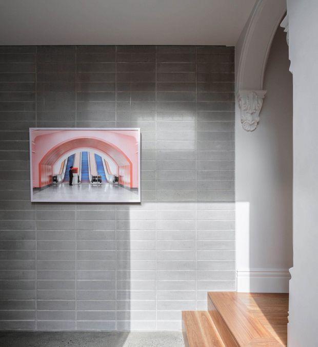 Interior view of a stair hall featuring a grey tiled feature wall, warm timber stairs, white painted walls and ornate decorative arch trim. A framed architectural artwork hangs on the tiled wall, with strong sunlight creating a bright geometric patch across the surfaces.