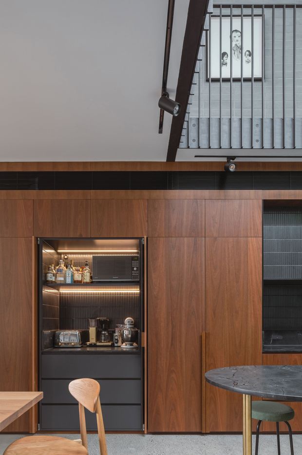 A sophisticated contemporary kitchen and dining area with floor-to-ceiling dark timber joinery, a concealed illuminated bar and coffee station, dark stone benchtops, and integrated appliances. A round marble-look dining table with a green upholstered chair occupies the foreground. The room features a high ceiling with a dramatic dark structural beam, track lighting, and a metal balustrade overlooking an upper level or stair void. The palette is warm timber, charcoal, black, and muted green, creating a refined, moody atmosphere.