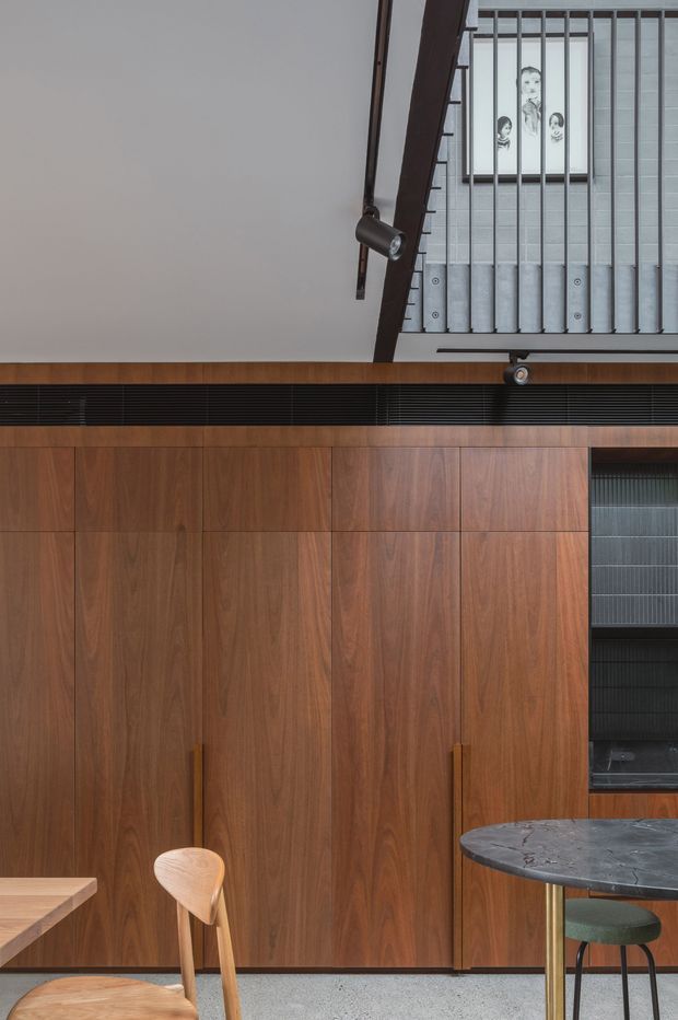 A refined interior corner featuring full-height dark timber wall panelling and concealed cabinetry, a black linear ventilation grille, and a small round dark stone-topped table with a green upholstered chair. A timber chair and partial dining surface appear in the foreground. The space has a restrained contemporary character with exposed vertical metal balustrades and a framed artwork visible on an upper level.
