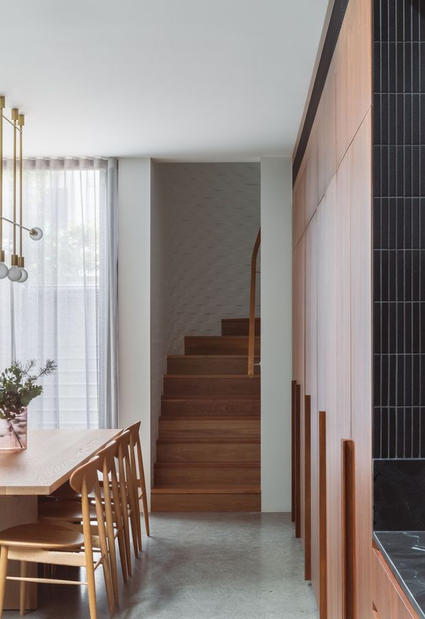 A bright, refined open-plan dining and circulation area with a timber dining table and spindle-back chairs beside a tall sheer-curtained window. A compact timber staircase with a curved handrail leads upward at the centre, while full-height timber cabinetry and a dark vertically tiled feature wall line the right side. The space combines warm timber, pale walls, polished concrete flooring and abundant diffuse daylight.