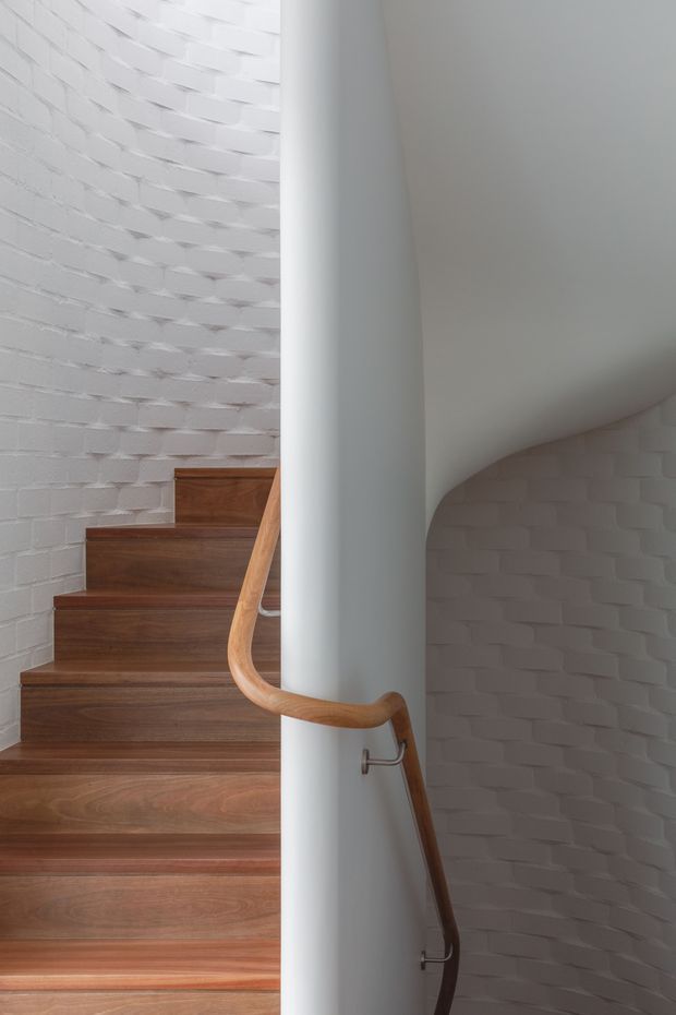 A curved interior stairway with warm timber treads and a smooth timber handrail wrapping around a central white structural column. The surrounding walls are white painted brick with a subtly textured, curved form, creating a sculptural and minimalist architectural composition.