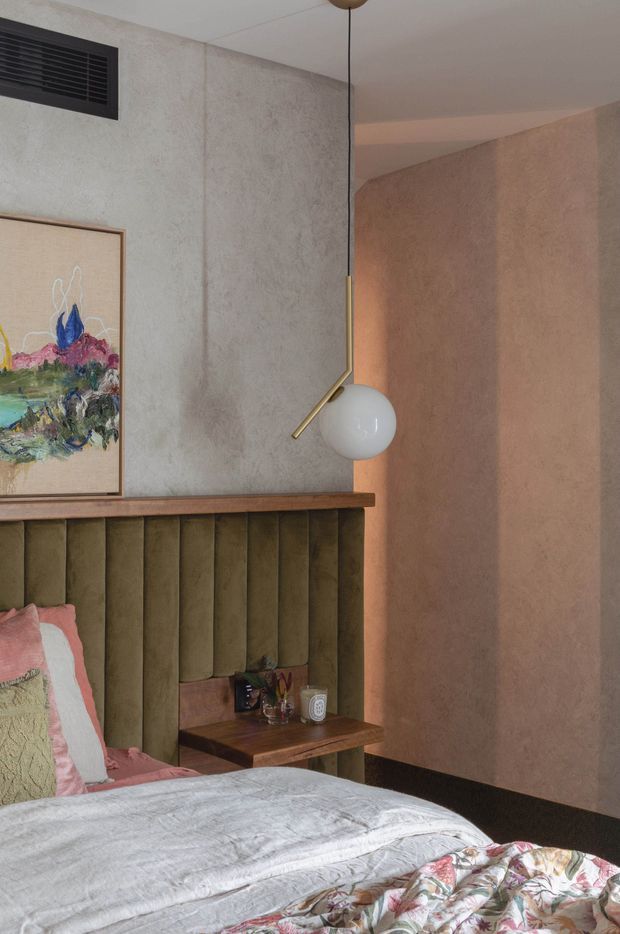 A warmly styled bedroom corner featuring a vertically channelled olive-green upholstered bedhead with timber ledge, a compact dark timber bedside table, layered pastel bedding, textured plaster walls, framed artwork, and a sculptural brass-and-white globe pendant light. The space has a low, intimate ceiling and softly atmospheric lighting.