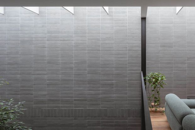 A close architectural view of a dark grey tiled feature wall enclosing a small planted courtyard or terrace. Horizontal rectangular tiles create a strong linear pattern, with white structural roof elements and narrow skylight openings above. Timber decking, potted greenery, and the edge of a curved upholstered sofa are visible at the right, suggesting an outdoor lounge or semi-enclosed courtyard setting.
