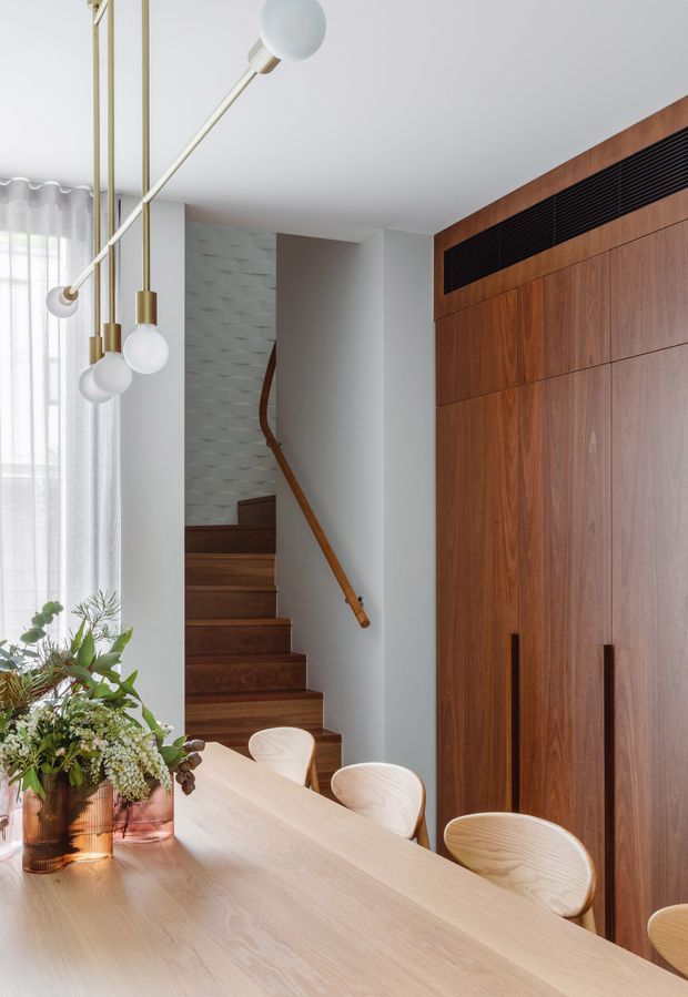 A refined dining area with a large pale timber dining table and curved light-wood chairs, opening onto a timber staircase. The room features a full-height dark timber built-in wall with black horizontal ventilation detailing, a sculptural multi-globe pendant light, sheer curtains, and indoor greenery. The palette is warm timber, white, and soft neutral tones with a clean contemporary feel.