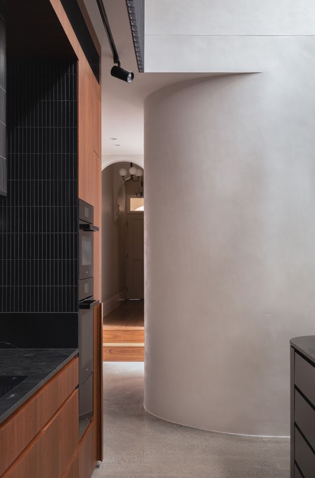 A refined interior view from a compact kitchen toward a hallway and stepped adjoining space. The kitchen features warm vertical timber cabinetry, a dark charcoal tiled splashback, dark stone benchtops, integrated black appliances, and black track lighting. A large softly curved plastered wall dominates the right side, contrasting with the linear joinery. The passage beyond has timber flooring, an arched opening, and sculptural pendant lighting. The palette is warm, muted, and minimalist with strong curved-versus-linear architectural detailing.