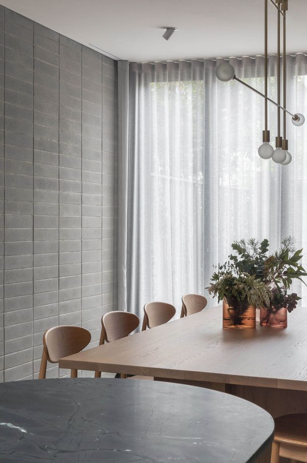 A refined dining area with a long light-wood dining table and sculptural wooden chairs, set beside a full-height window dressed with sheer grey curtains. The room features a grey block-pattern masonry wall, a contemporary multi-arm pendant light, and warm-toned botanical arrangements in copper vessels. A dark stone-topped counter or island occupies the foreground.