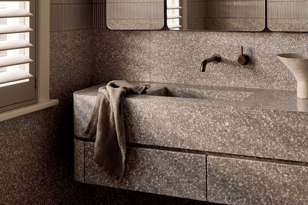 Close-up view of a refined bathroom vanity with an integrated rectangular basin, wall-mounted bronze tapware, terrazzo-clad benchtop and splashback, and matching floating drawer cabinetry. A louvered window sits to the left, with a textured towel draped over the basin edge and a sculptural vessel on the right. The palette is warm and muted, with strong tonal continuity and tactile stone-like surfaces.