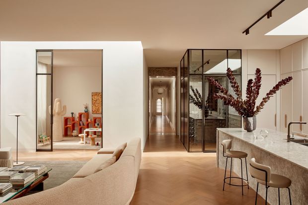 A refined open-plan contemporary interior showing a living room, kitchen and central circulation corridor. A curved cream sofa faces a low coffee table and floor lamp, while the kitchen features a pale stone island with integrated sink, dark faucet and bar seating. Full-height black-framed glazed partitions enclose a secondary room beside the hallway, and warm timber flooring runs throughout. The palette is warm and neutral with sculptural floral branches and colourful objects providing accents.