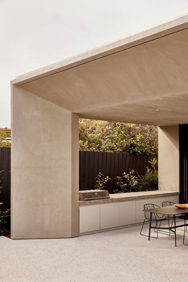 A refined covered outdoor entertaining area with a large rendered masonry canopy and substantial square columns. The space includes a built-in outdoor kitchen with pale cabinetry, stone-look benchtop and integrated barbecue, opening onto a landscaped courtyard with dark fencing. A compact dining table and wire-frame chairs sit on a light speckled paved or terrazzo-like floor. The composition is warm, minimal and contemporary, with strong geometric lines and sheltered indoor-outdoor character.