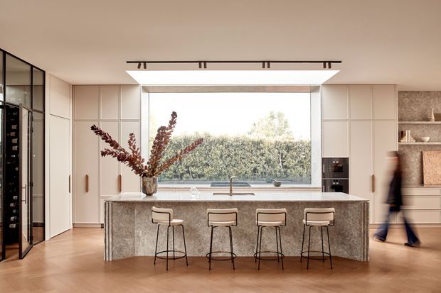 A refined contemporary kitchen viewed straight on, centred around a large stone island with four slim bar stools. Full-height light cabinetry frames a broad picture window above the sink, while a wine display or refrigerator enclosure is visible at left and open shelving with integrated cabinetry appears at right. Warm timber flooring, pale cabinetry, a textured stone island and linear ceiling lighting create a calm, polished interior.